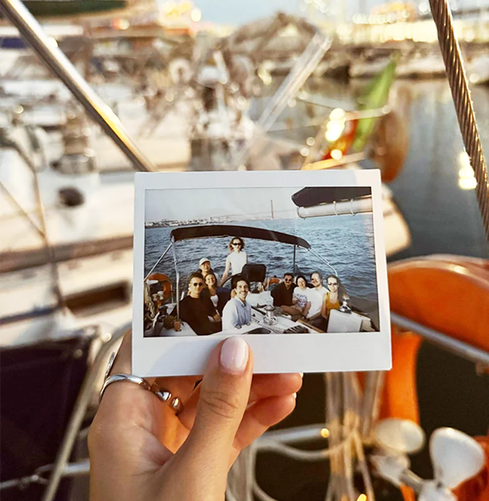 A hand holding a Polaroid of the Phira team gathered on a sailing boat at sunset, taken in the marina behind it